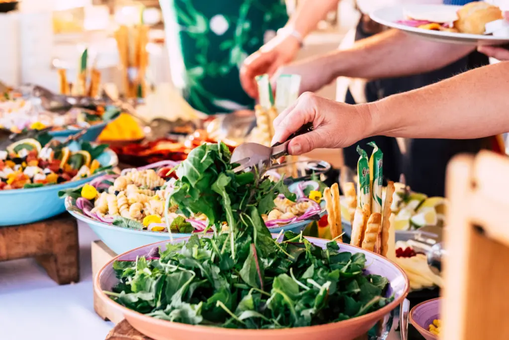 A buffet table filled with healthy salads and baked dishes with the hands of people dishing up the food onto plates.